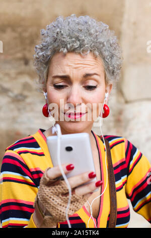 Ritratto di trafitto donna matura con il grigio capelli ricci guardando al telefono cellulare Foto Stock