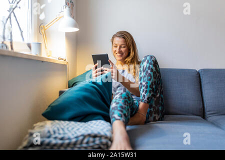 Felice giovane donna seduta sul divano di casa utilizzando tablet Foto Stock