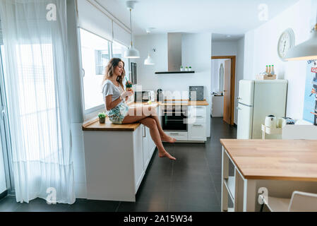 Giovane donna seduta sul banco di cucina con un drink Foto Stock