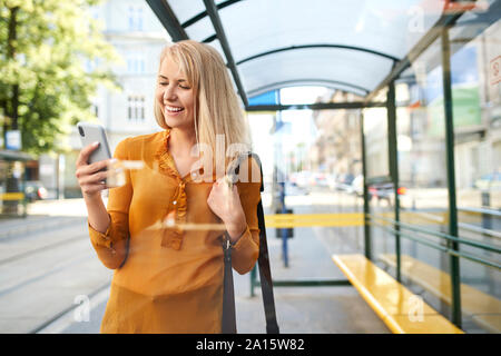 Sorridente giovane donna utilizza lo smartphone alla fermata del tram Foto Stock