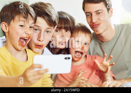 La madre e i suoi quattro figli prendendo selfies smartphone a pranzo, con facce piene di sugo di pomodoro Foto Stock