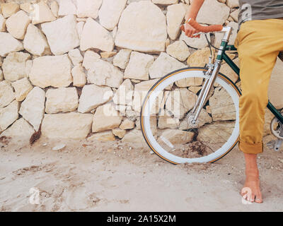 A piedi nudi uomo con Fixie bike davanti al muro di pietra naturale sulla spiaggia, vista parziale Foto Stock