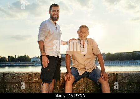 Ritratto di sorridere padre e figlio adulto in riva al fiume e al tramonto Foto Stock