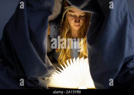 Giovane donna illuminata di lettura libro nel letto di casa Foto Stock