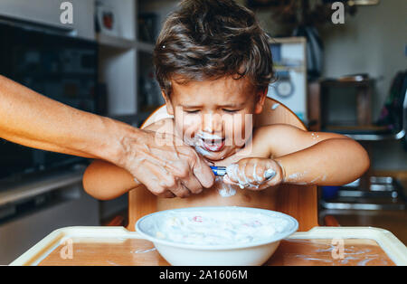 Little Boy mangiare lo yogurt a casa, mano della donna sul cucchiaio Foto Stock