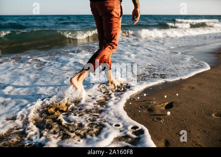 Vista posteriore dell'uomo con orange pantaloni camminando su una spiaggia a bordo d'acqua Foto Stock