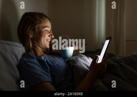 Giovane donna giaceva a letto a casa con la tazza di tè di notte utilizzando tablet Foto Stock