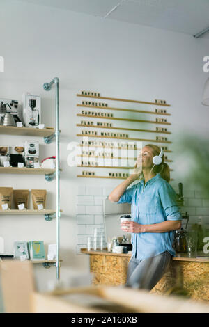 Giovane donna ascoltando musica, indossando le cuffie, stando in piedi in un coffee shop Foto Stock