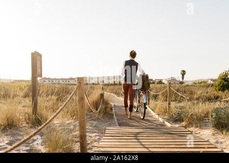 Ben vestito uomo a camminare con la sua bicicletta su una passerella in spiaggia al tramonto Foto Stock