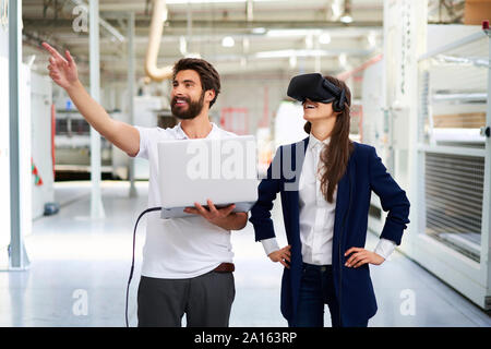 Uomo con notebook e imprenditrice indossando occhiali VR in fabbrica Foto Stock