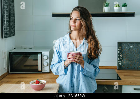 Ritratto di giovane donna che indossa pigiama in cucina a casa azienda tazza di caffè Foto Stock