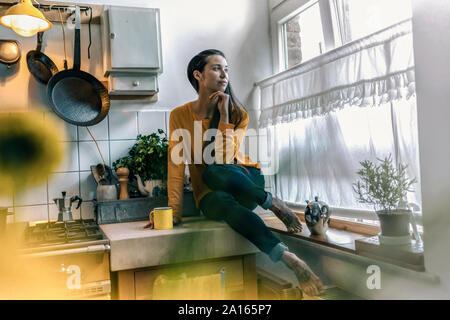Giovane donna seduta sul banco di cucina a casa guardando fuori della finestra Foto Stock