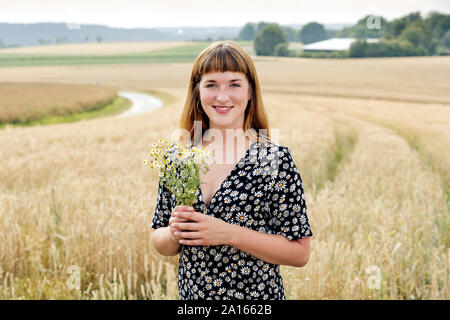 Ritratto di sorridente giovane donna con il mazzo di fiori di camomilla in piedi nella parte anteriore del campo di grano Foto Stock