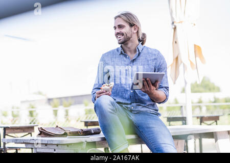 Giovane uomo seduto a tavola in un giardino della birra con il tablet e sandwich Foto Stock