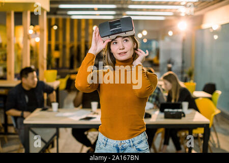 Bella femmina business donna in arancione maglione cercando 3d occhiali VR al lavoro. Multirazziale team di giovani che lavorano insieme in ufficio creativo Foto Stock