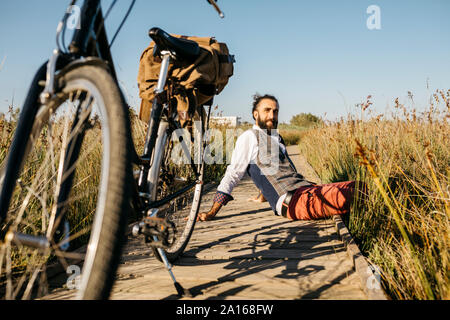 Ben vestito uomo seduto su una passerella di legno in campagna accanto a una bicicletta Foto Stock