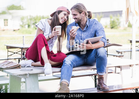 Coppia giovane seduto sulla tavola in un giardino della birra con la compressa, sandwich e caffè da asporto Foto Stock