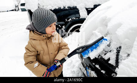 Closeup colpo di adorabile ragazzo felice nel rivestire aiutando a pulire la neve auto coperto con spazzola Foto Stock