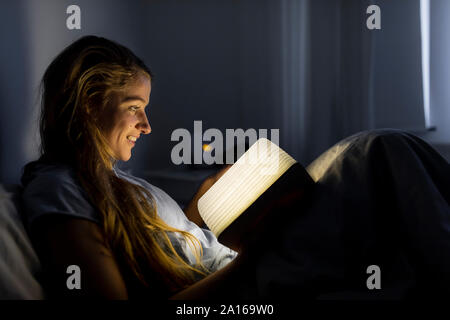 Sorridente giovane donna illuminata di lettura libro nel letto di casa Foto Stock