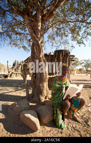 Tradizionale donna muhila, seduto sotto agli alberi, la lettura di un libro, Kehamba, Chibia, Angola Foto Stock