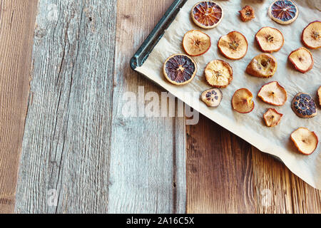 In casa delle fette di essiccato le arance e le mele sulla carta da forno e nero vassoio da forno, disposizione diagonale dall'angolo superiore destro, sullo sfondo di legno Foto Stock