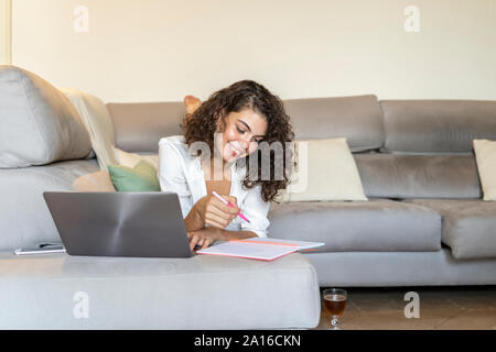 Sorridente giovane donna prendendo appunti e utilizzando laptop sul lettino a casa Foto Stock