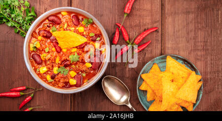 Un panorama del cibo messicano. Chili con carne, peperoncino, foglie di coriandolo, e nachos, girato dalla parte superiore al buio su un rustico sfondo di legno Foto Stock