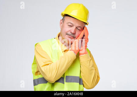 Senior repairman ispanica nel casco giallo è stanco e assonnato dopo la dura giornata lavorativa. Ho bisogno di un concetto di riposo. Studio shot Foto Stock