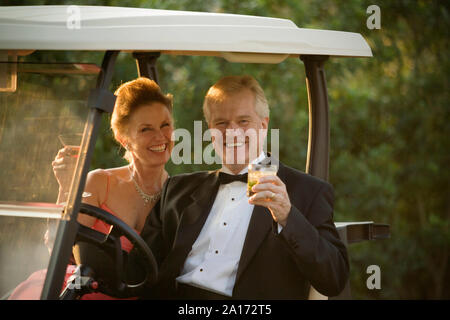Ritratto di una coppia matura vestito formalmente seduto su un carrello da golf con cocktail. Foto Stock