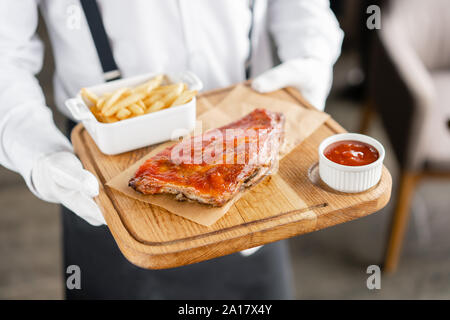 Il cameriere è in possesso di un piatto di legno deliziose costolette di maiale. Rack completo di nervature barbeque sul piatto di legno con patatine fritte e insalata mista Foto Stock