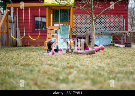 Due bambini felici laici sulle stuoie nel loro cortile chiacchierare insieme Foto Stock