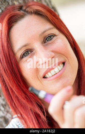 Ritratto di una bella donna redhead appoggiata contro un albero di fumare una sigaretta elettronica in un parco Foto Stock