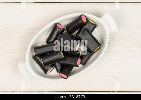 Sacco di tutto il dolce di liquirizia nera candy in bianco ovale tazza ceramica flatlay su legno bianco Foto Stock