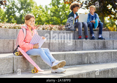 Ritratto di ragazza seduta sulle scale con lo skateboard e sorridente in telecamera durante la lettura di un libro con i suoi compagni in background Foto Stock