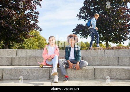 Ritratto di bambina con African ragazzo seduto su scale e riposo dopo lo skateboard nel parco Foto Stock