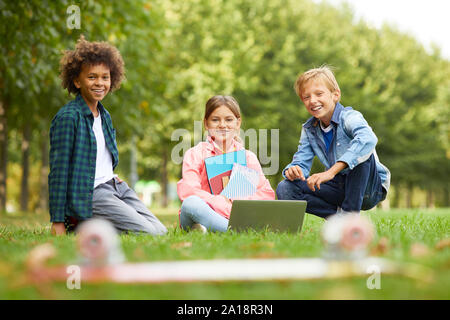 Ritratto di gruppo multietnico di amici seduti sul prato verde con libri e computer laptop e sorridente alla videocamera all'esterno Foto Stock
