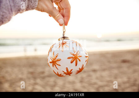 Donna bianca tenendo palla di Natale sulla spiaggia al tramonto in California, stagione Concetto di vacanza - Immagine Foto Stock