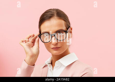 Close-up di giovane donna gravi in occhiali lavora come insegnante in posa in telecamera su sfondo rosa Foto Stock