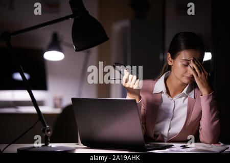 Giovane imprenditrice di toccare la sua testa mentre è seduto al suo posto di lavoro con computer portatile ella stanco del suo lavoro in ufficio Foto Stock