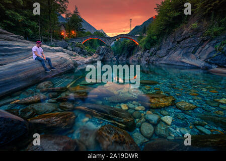 Turista gode il tramonto in un fiume vicino al ponte in pietra Lavertezzo, Svizzera Foto Stock