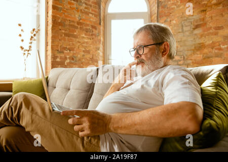 Senior uomo che lavora con il portatile a casa - Concetto di studiare a casa. Caucasica modello maschile seduto sul divano e facendo il suo dovere mentre serfing in internet, guardare il cinema o webinar, lezioni online. Foto Stock