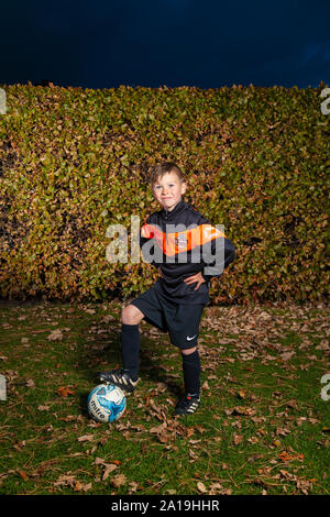 Un bambino di otto anni che indossa una striscia di calcio con le braccia sui fianchi e il suo piede su un campo di calcio Foto Stock