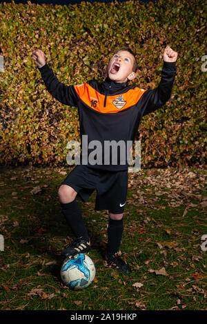 Un bambino di otto anni che indossa una striscia di calcio con le braccia sollevate e gridando. Foto Stock