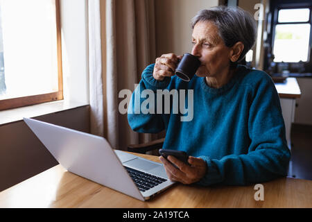 Senior donna a bere caffè e utilizza lo smartphone a casa Foto Stock