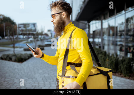 Giovani corriere a consegnare il cibo con borsa termica, controllo dell'ordine su uno smart phone mentre in piedi sulla strada della citta'. Consegna sul concetto del piede Foto Stock