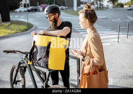 Corriere maschio a consegnare il cibo con una bicicletta a un giovane imprenditrice, ottenere alcuni pacchetti da una busta termica all'aperto Foto Stock