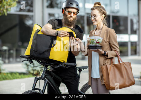 Corriere consegna pranzi freschi ad una giovane donna di affari su una bicicletta con zaino termico. Il ristorante da asporto cibo concetto di consegna Foto Stock