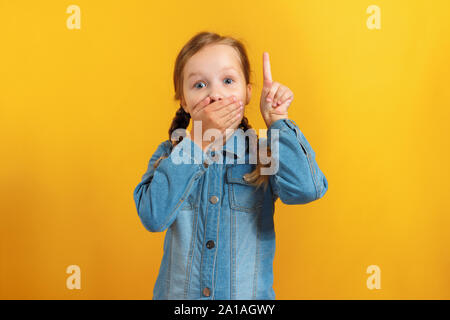 Bambina su uno sfondo giallo. Il bambino è stupito chiudendo la bocca con la mano e indicando con un dito indice per un posto vuoto. Foto Stock