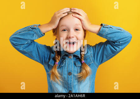 Bambina tiene le mani dietro la testa su uno sfondo giallo. Il bambino è stupito, sorpreso, emotiva. Foto Stock