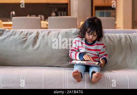 Piccola ragazza seduta sul suo divano utilizzando una tavoletta digitale Foto Stock